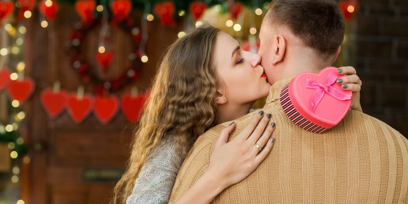 woman giving a kiss to a man holding a Valentine's Day gift box in front of a Valentine's Day celebration and decorations