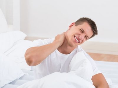 Young Man Suffering From stiff neck In Bedroom