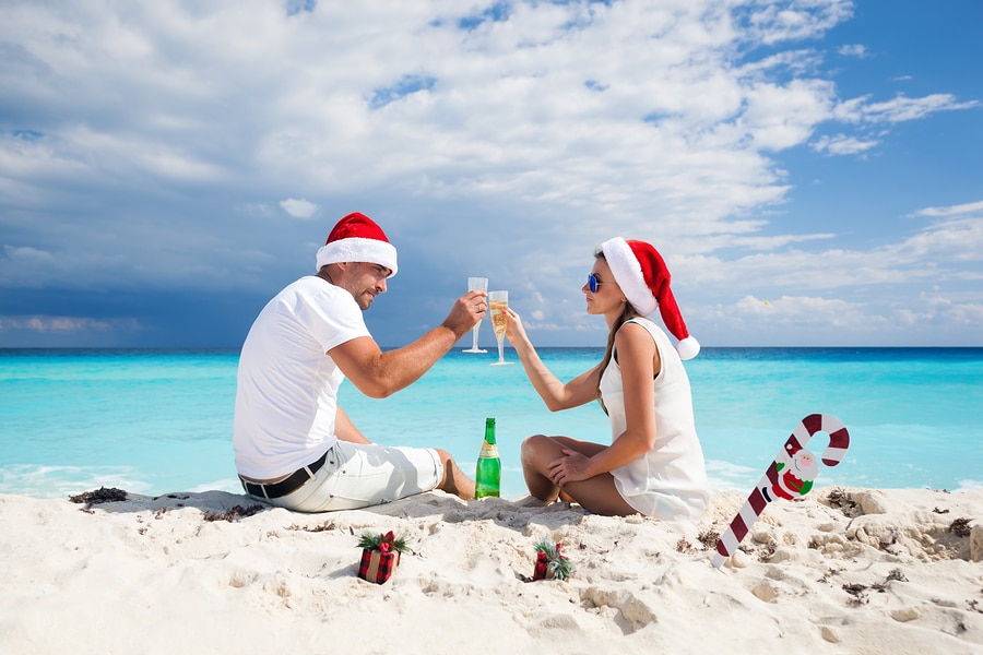 Happy couple in red Santa hats celebrating Christmas on beach drinking champagne