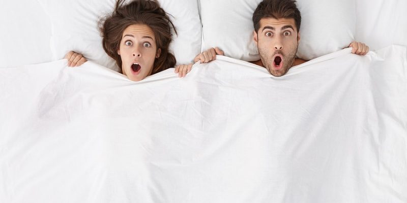 man and woman looking surprised in bed on white pillows