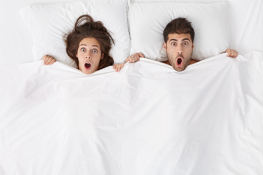 man and woman looking surprised in bed on white pillows