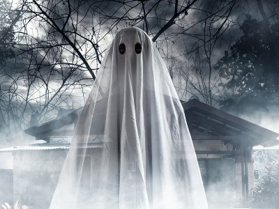 Halloween ghost in bed sheet costume in front of foggy haunted house and trees