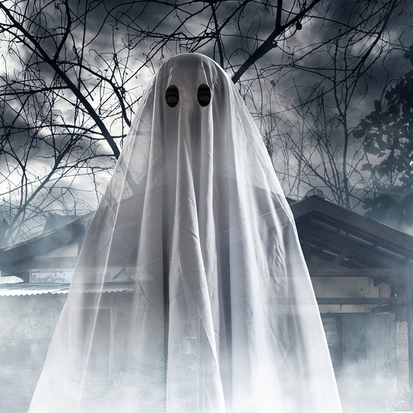 Halloween ghost in bed sheet costume in front of foggy haunted house and trees