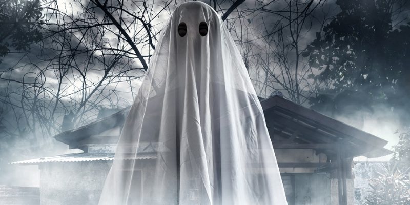 Halloween ghost in bed sheet costume in front of foggy haunted house and trees