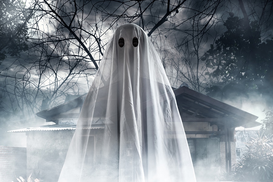 Halloween ghost in bed sheet costume in front of foggy haunted house and trees