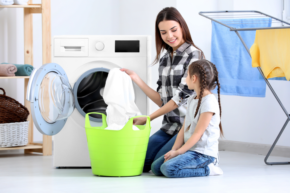 mom and young daughter loading laundry into washing machine sheets pillowcases and clothing