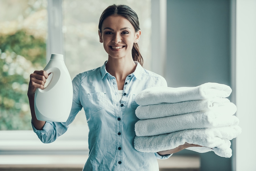 Young Smiling Woman holding Laundry Detergent. Happy Beautiful Girl holding Bottle of Loundry Detergent and fluffy clean towels properly laundered