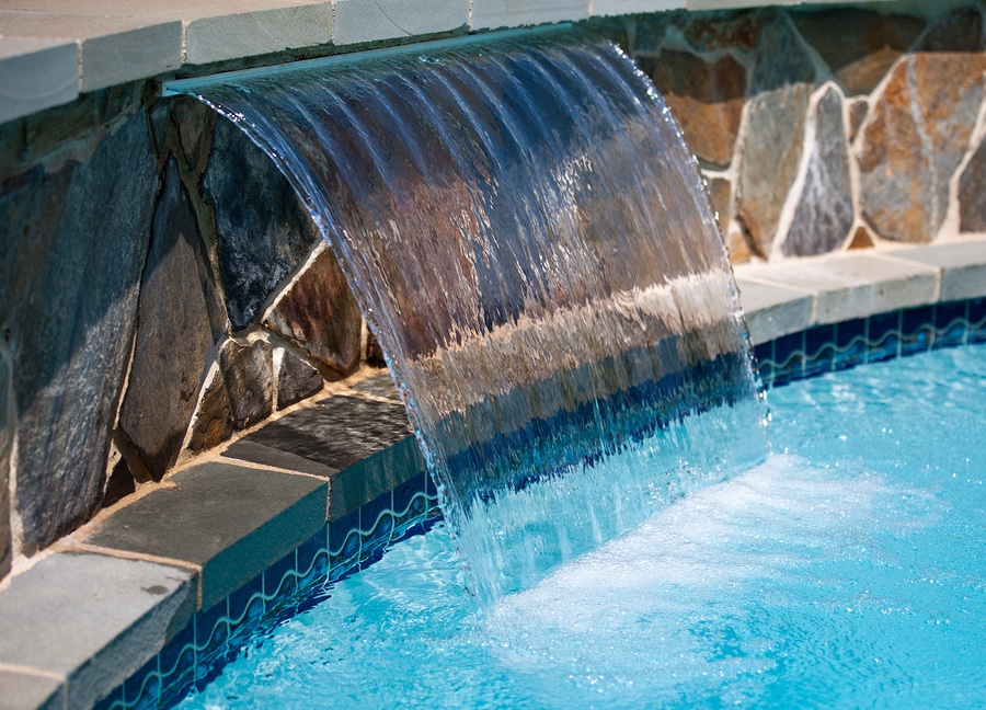 Plume of water arching out of a stone wall into a blue swimming pool