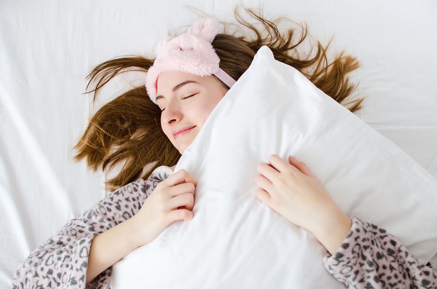 happy young woman in morning hugging pillow and wearing pajamas and bunny eyeshades for better sleep