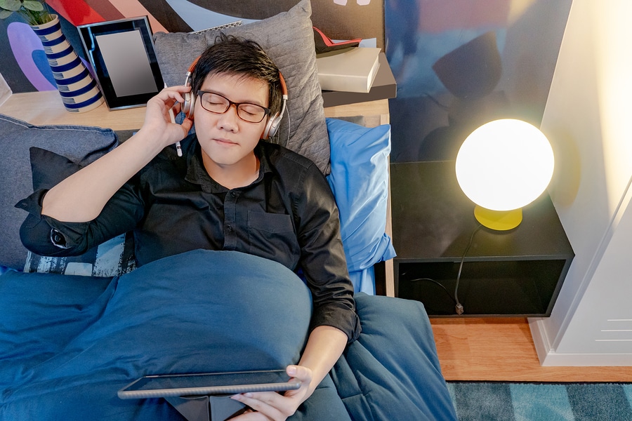 Young asian man in bed wearing headphones and holding tablet PC showing electronics used at bedtime