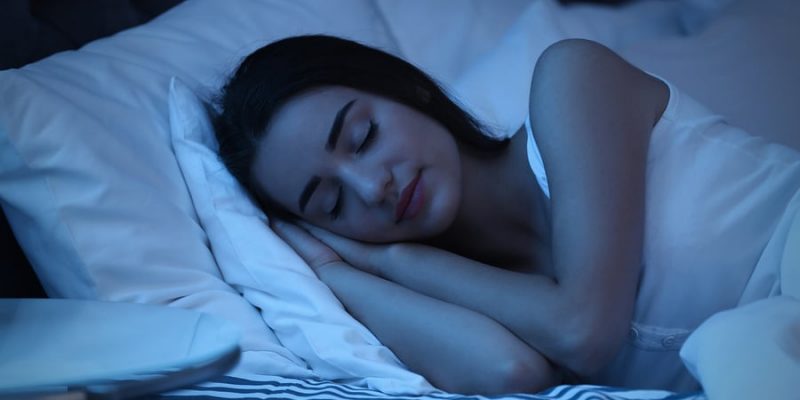 Relaxed smiling young woman sleeping in bed on two soft pillows in dark bedroom