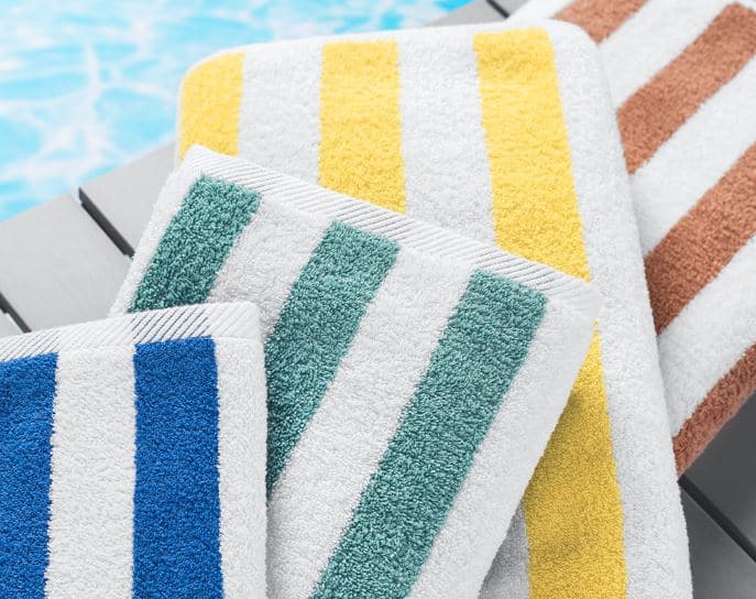 Sobel Westex Splash Stripe hotel pool towels in four colors on bench next to blue swimming pool
