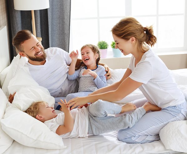 happy family of parents and two kids playing on summer bedding white bed sheets in sunny bedroom
