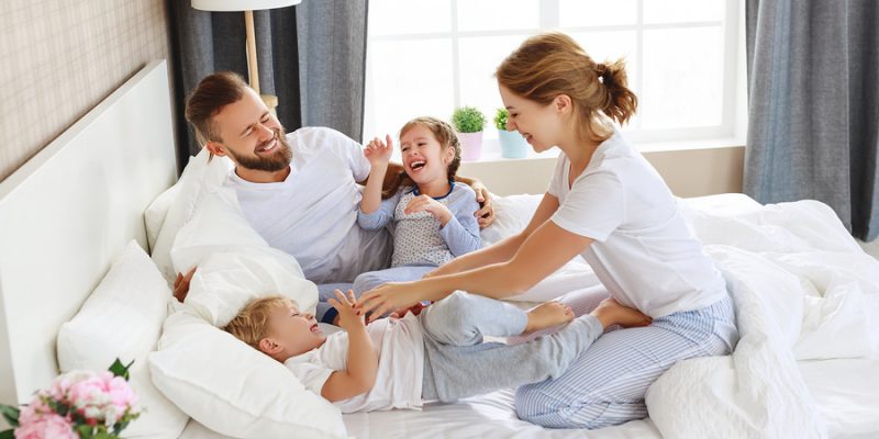 happy family of parents and two kids playing on summer bedding white bed sheets in sunny bedroom
