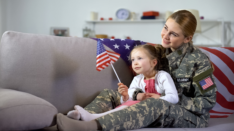 American female service member and little daughter at home on the sofa with flag watching armed services celebration