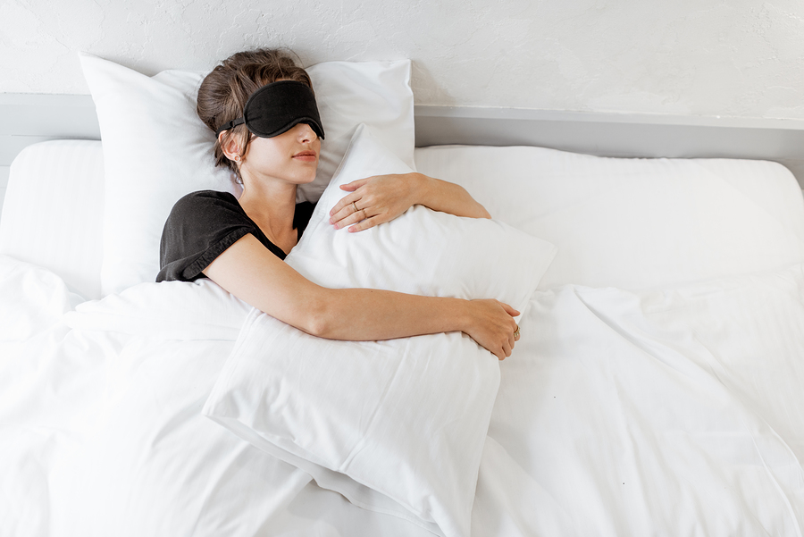 Woman lying with a sleeping mask in bed covered with sanitized white bed sheets in bright bedroom in the morning