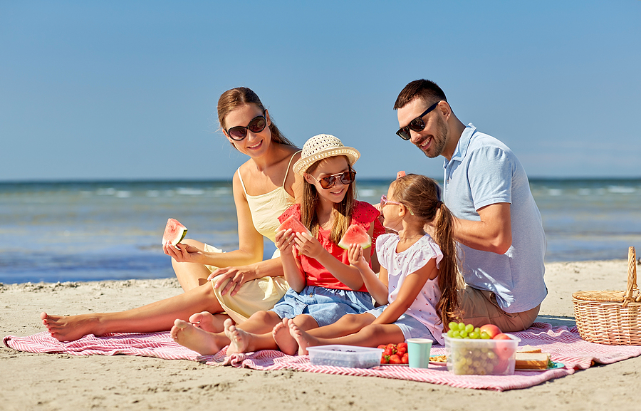 family of four on beach towel wiht picnic basket eating watermelon