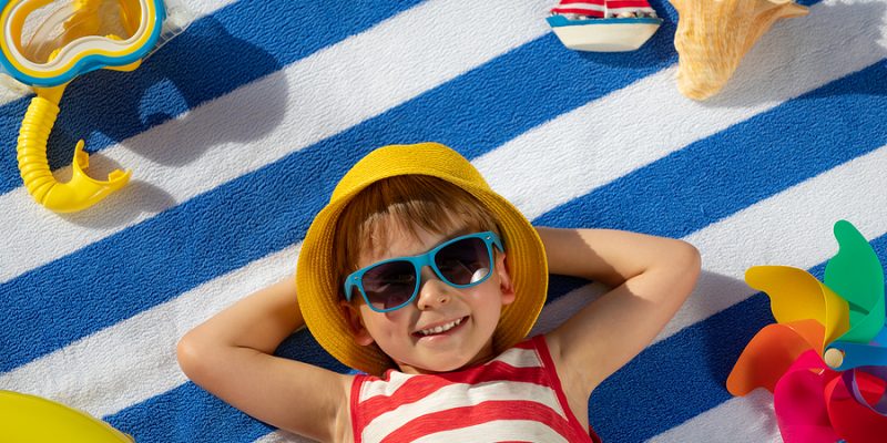 Happy small boy lying on blue and white striped cotton beach towel with beach toys around him