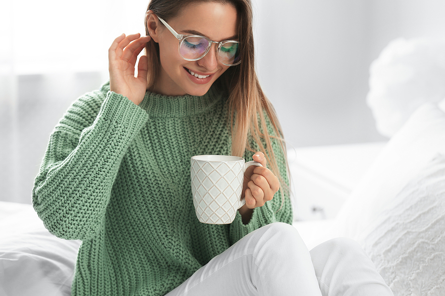Young woman in warm sweater with hot drink sitting on a white duvet covered bed