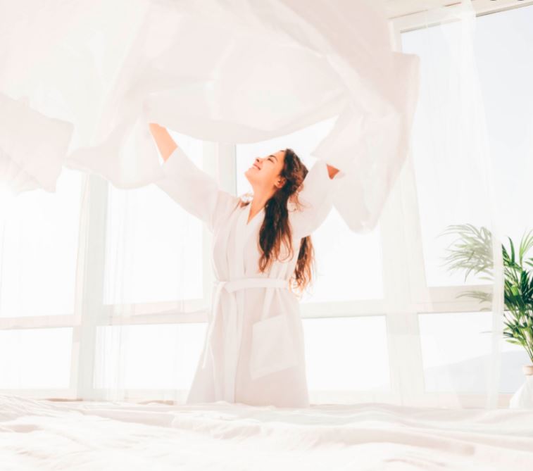 Happy woman in white robe making the bed tossing a white cotton sheet in bright sunny room with floor to ceiling windows