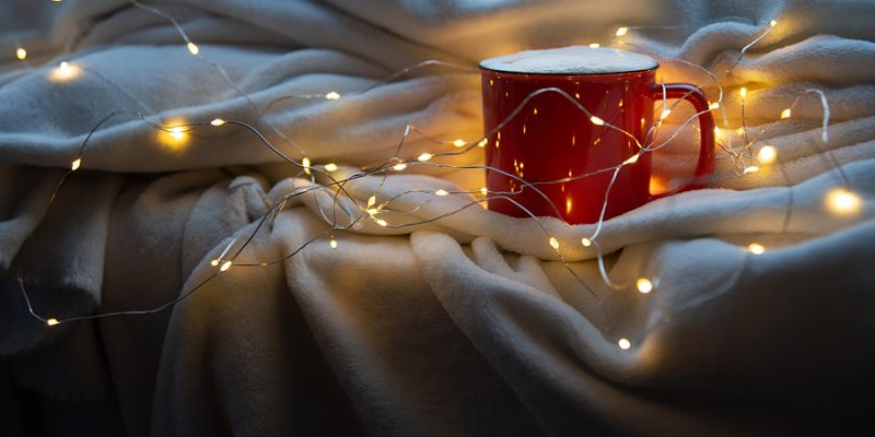 Holiday image of red and white mug of coffee or cocoa on white plaid with christmas lights.