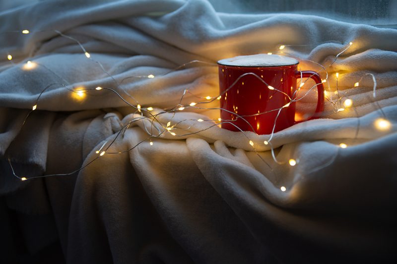 Holiday image of red and white mug of coffee or cocoa on white plaid with christmas lights.