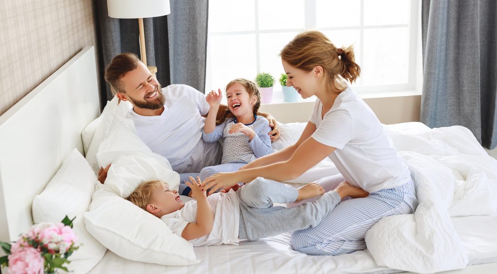 happy family mother, father and children laughing, playing and tickles in bed with fluffy down pillows