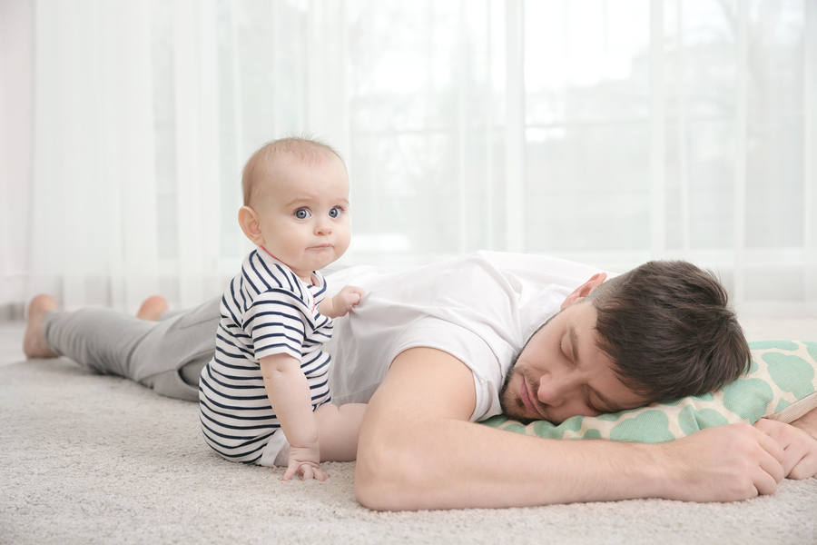 Tired dad sleeping on the floor on his stomach with a pillow, while baby tugs on his shirt.