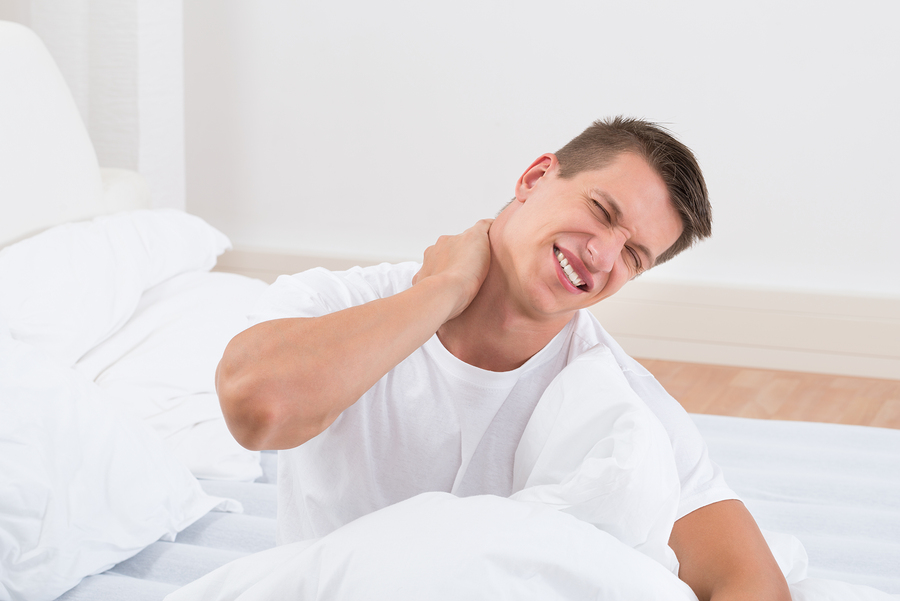 Man sitting up in bed, grimacing in pain from sleeping wrong or on an unsupportive pillow.
